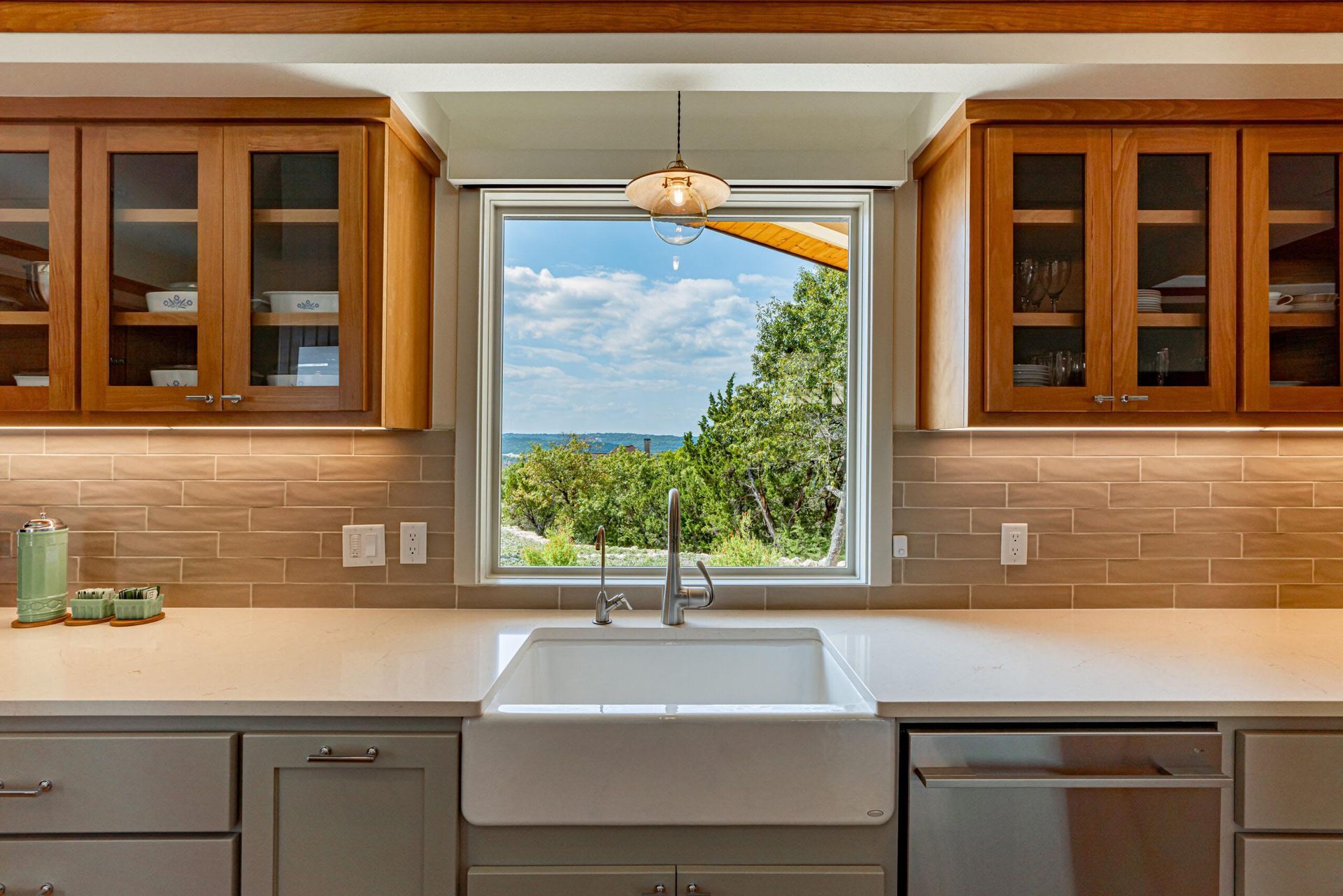 Kitchen with a white sink centered under a window offering a view of greenery and blue sky. Wooden cabinets.