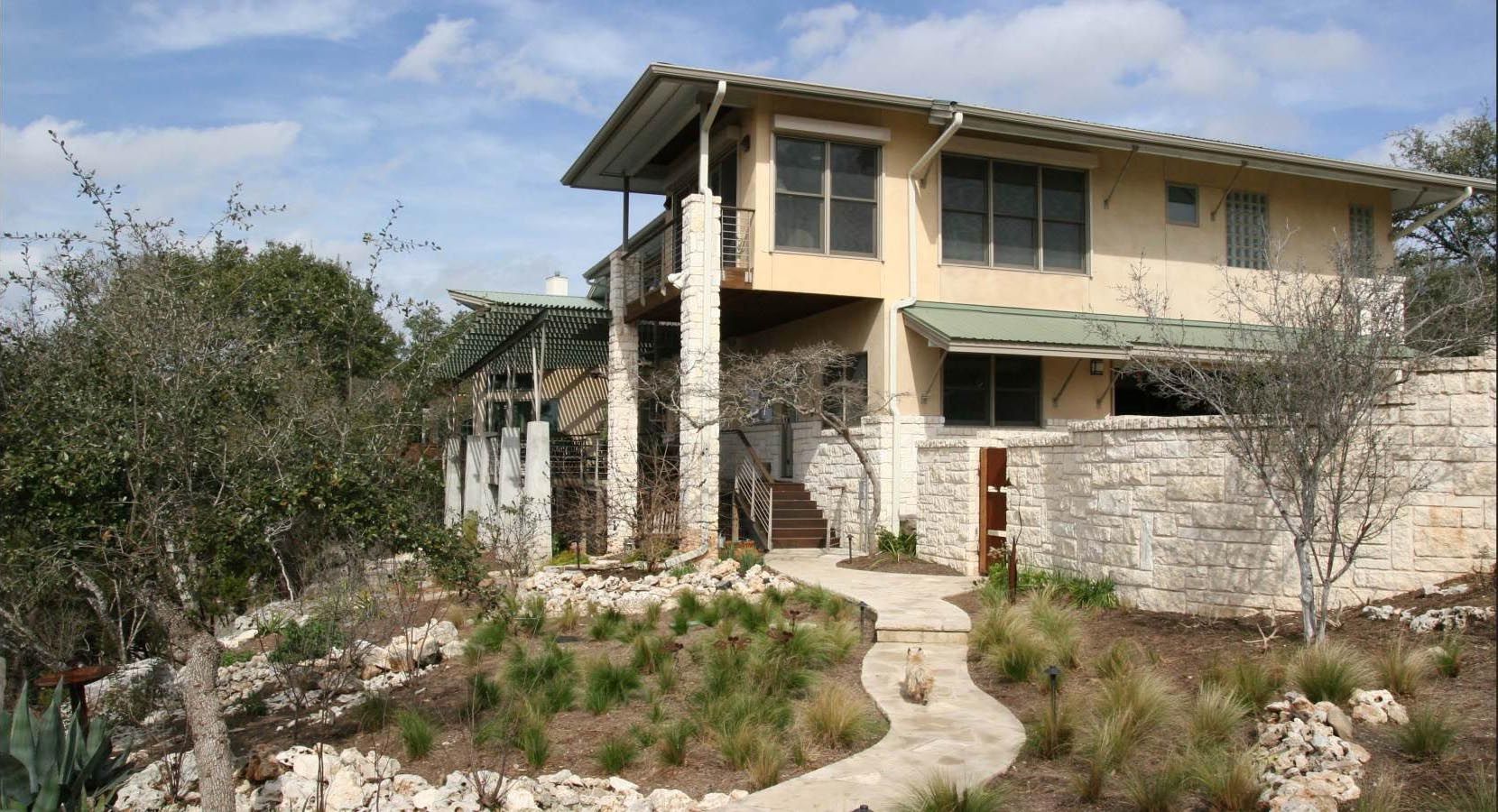 Two-story house with stone and stucco facade, pathway leads to front door, trees and shrubs surround.