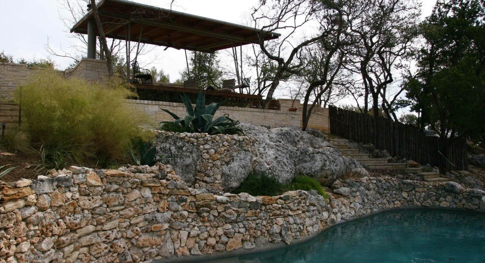 Stone retaining walls and steps lead to a covered patio above a pool.