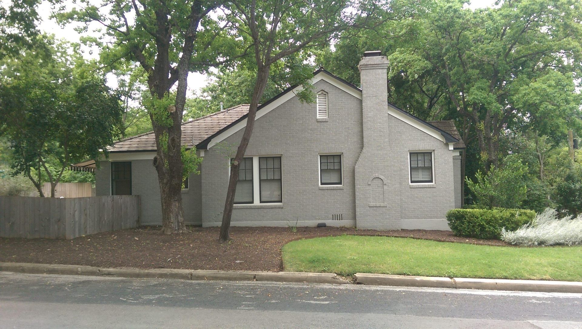 Gray brick house with dark roof, chimney, and two windows. Trees and a small lawn in front.