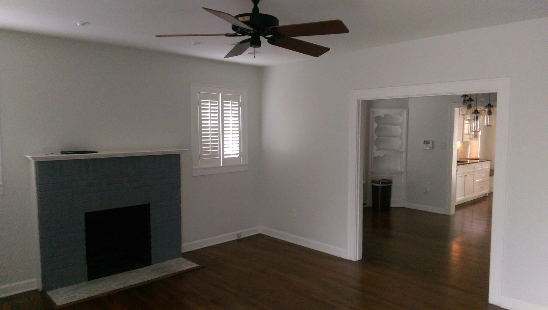 Living room with fireplace, window with shutters, and doorway to kitchen; dark wood floor, light walls.