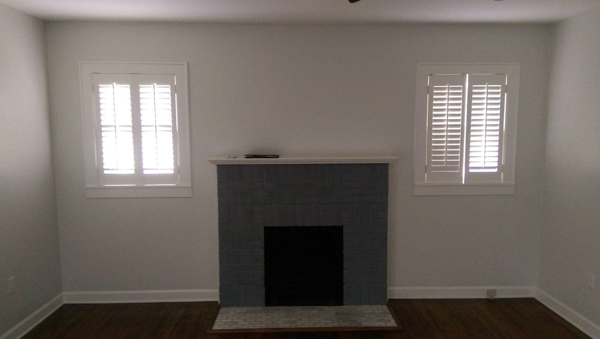 Living room with fireplace between two shuttered windows. Gray walls, fireplace, and hardwood floors.