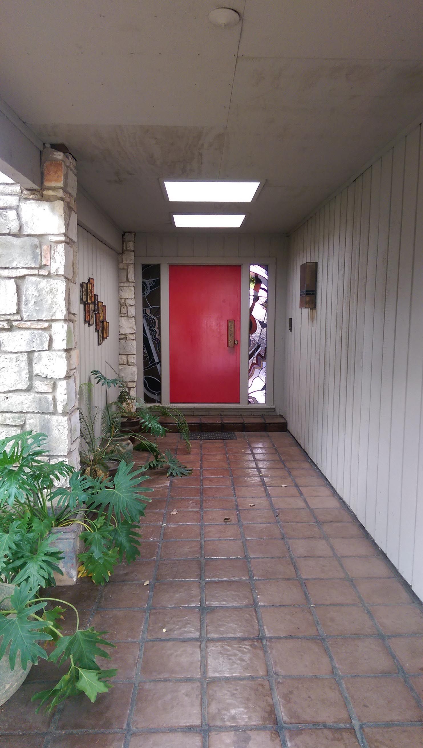 Red front door framed by glass, on a covered walkway. Brick floor, stone wall on left, and plants.