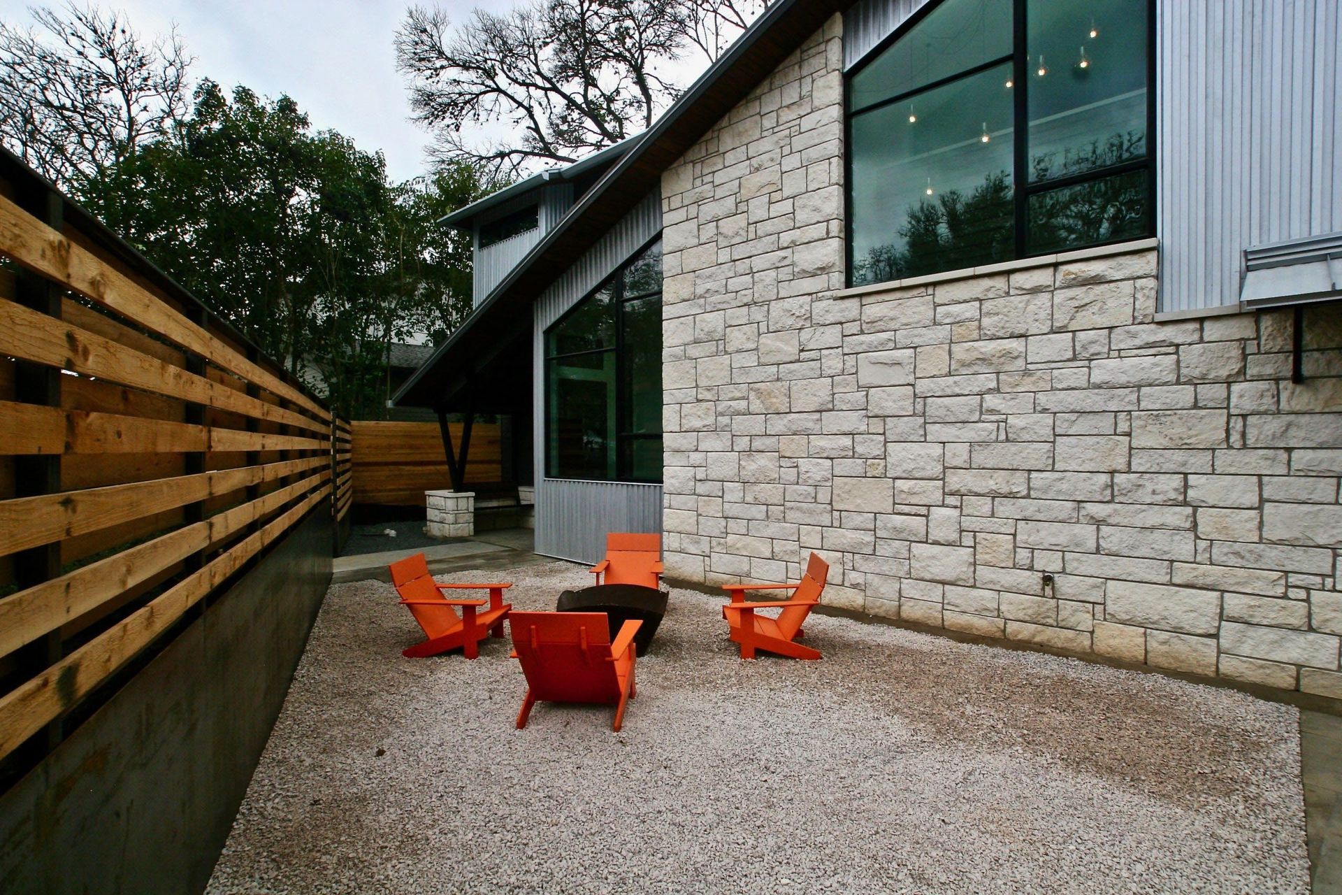 Outdoor patio with orange chairs, gravel ground, and stone wall. Wooden fence on the left.