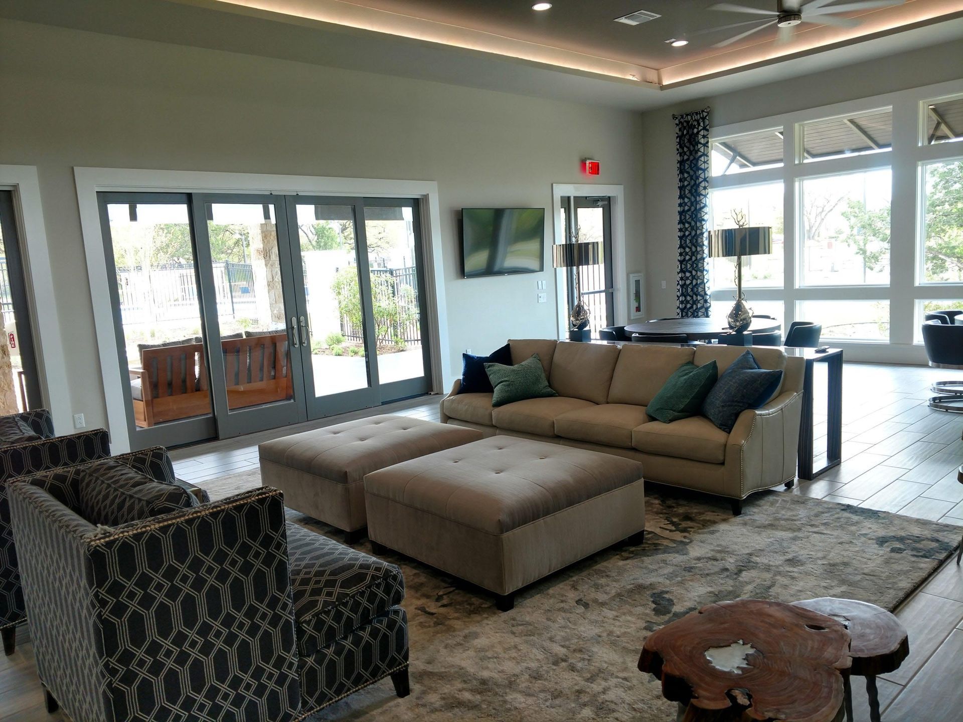 Spacious, well-lit modern living room with sliding glass doors, tan couch, patterned armchairs, and two ottoman benches.