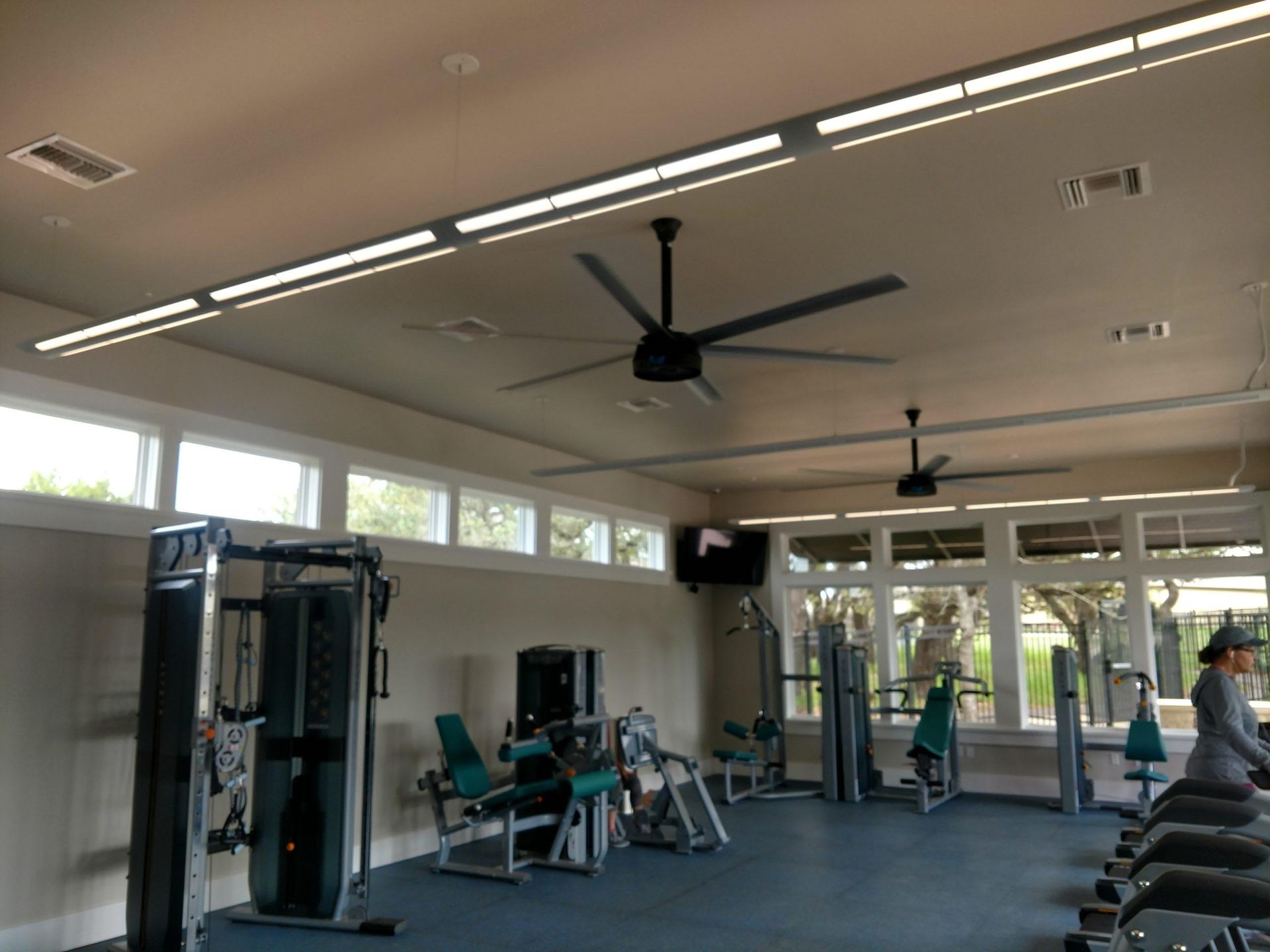 Gym interior with exercise equipment and ceiling fans. A person is on a treadmill near large windows.