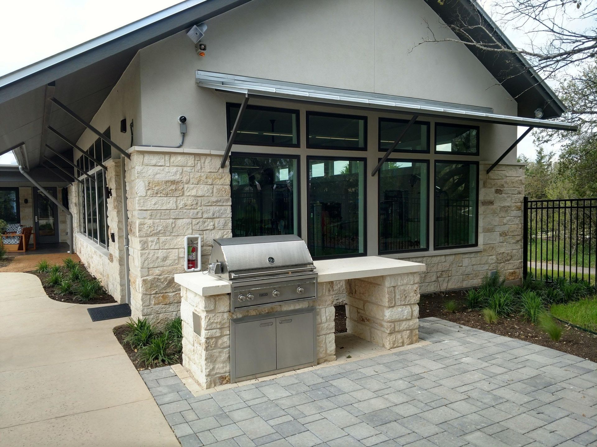 Outdoor kitchen with a stainless steel grill on a stone countertop under a building with large windows.