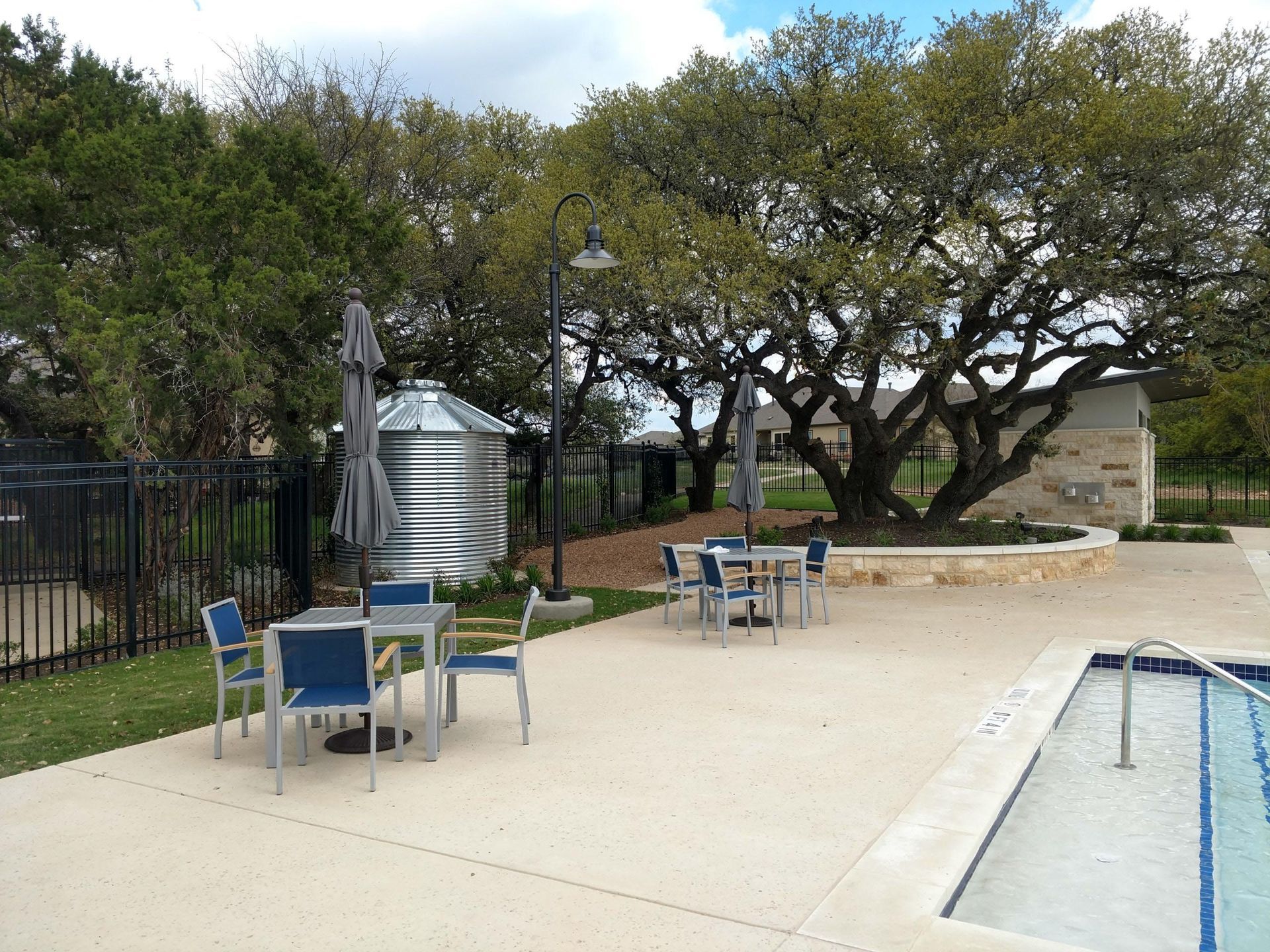 Tables and chairs near a pool under trees and an umbrella. Silo and building in background.