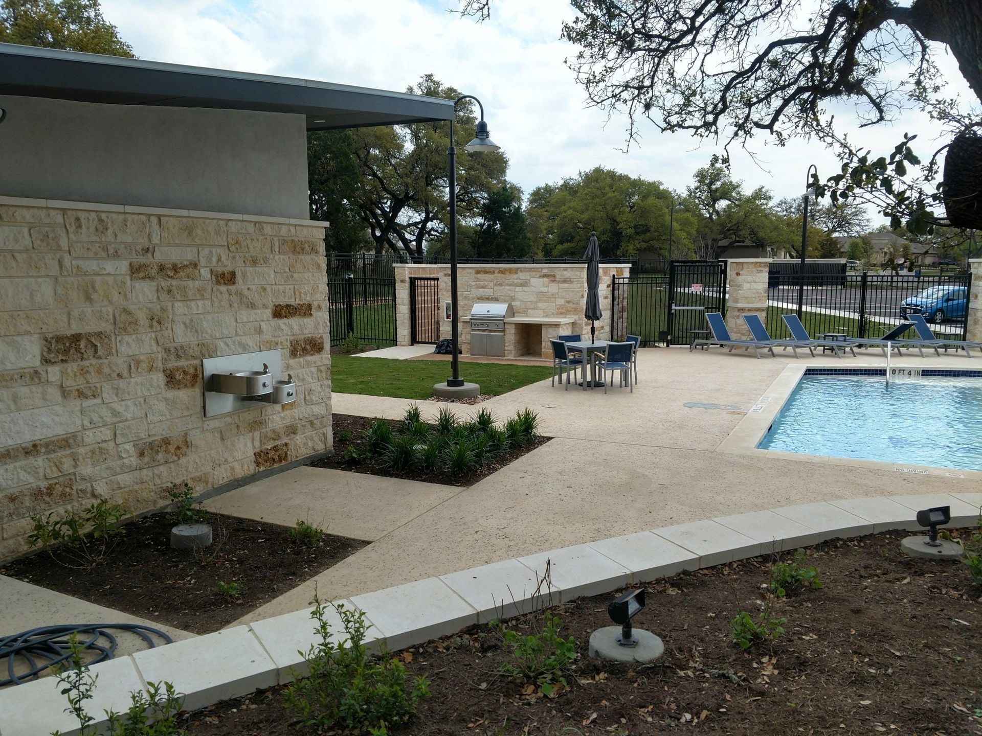 Poolside area with a grill, dining table, and lounge chairs. Beige stone building, blue pool.