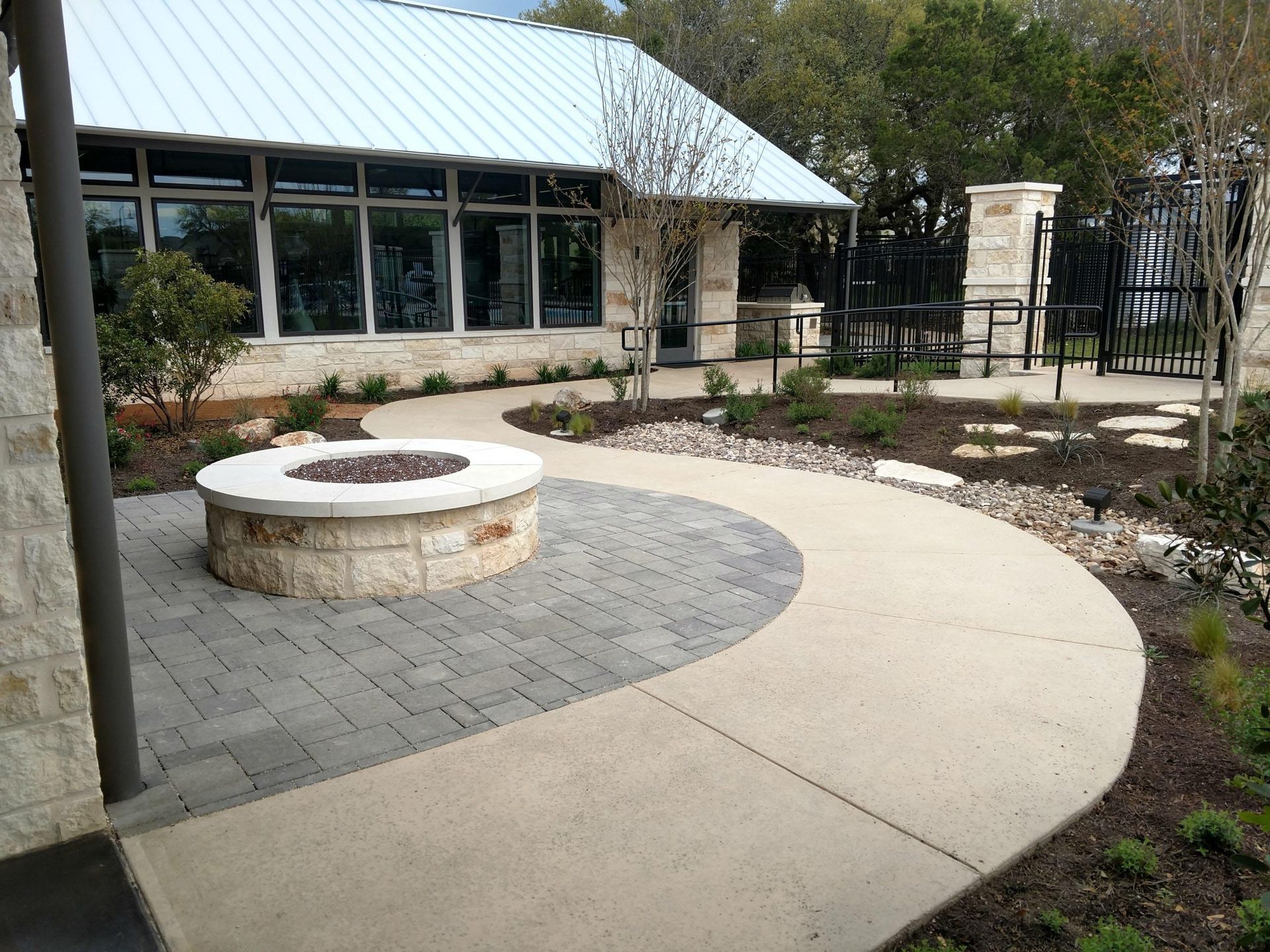 Stone fire pit on a paved patio with a curving walkway leading to a building with a metal roof.
