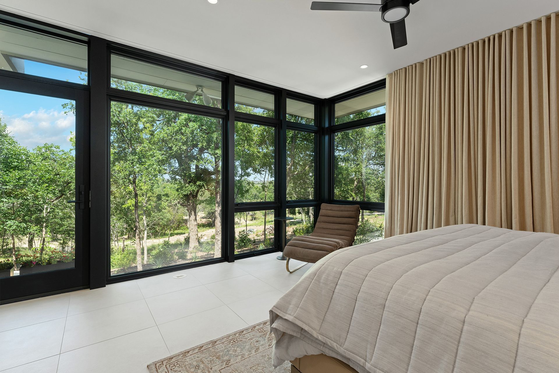 Bedroom with large windows, trees visible outside. Light beige bed, lounge chair, and curtains.