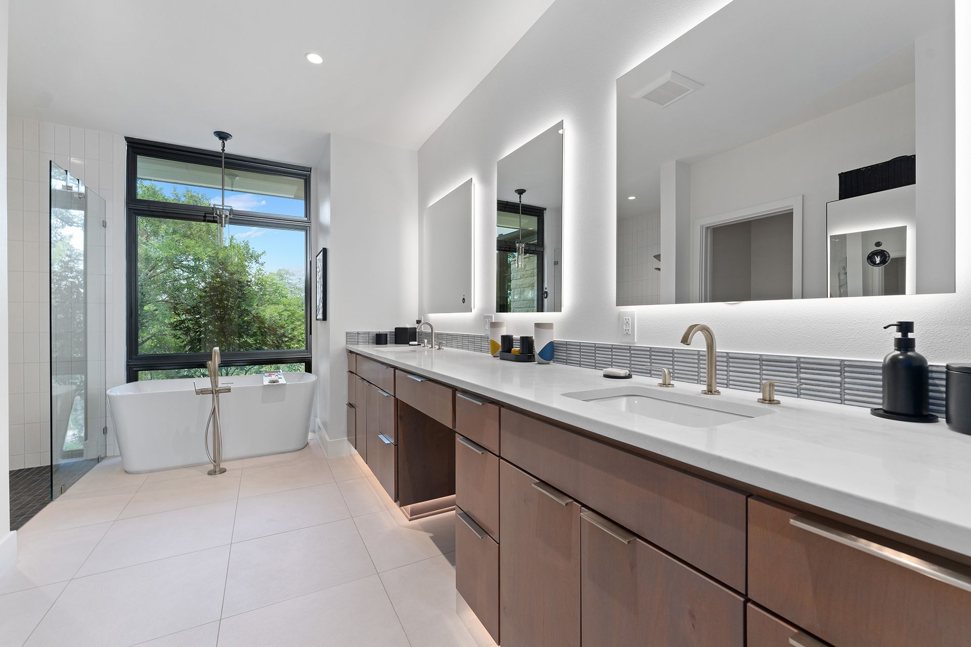 Modern bathroom with a long vanity, large mirrors, a window with a tub, and wood cabinetry.
