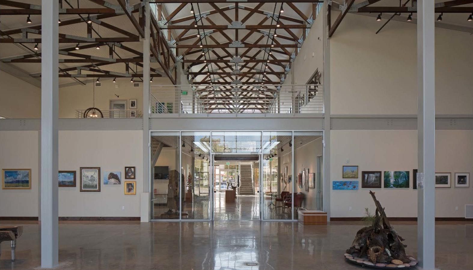 Art gallery interior with white pillars, glass walls, and artwork. A wooden structure is visible overhead.