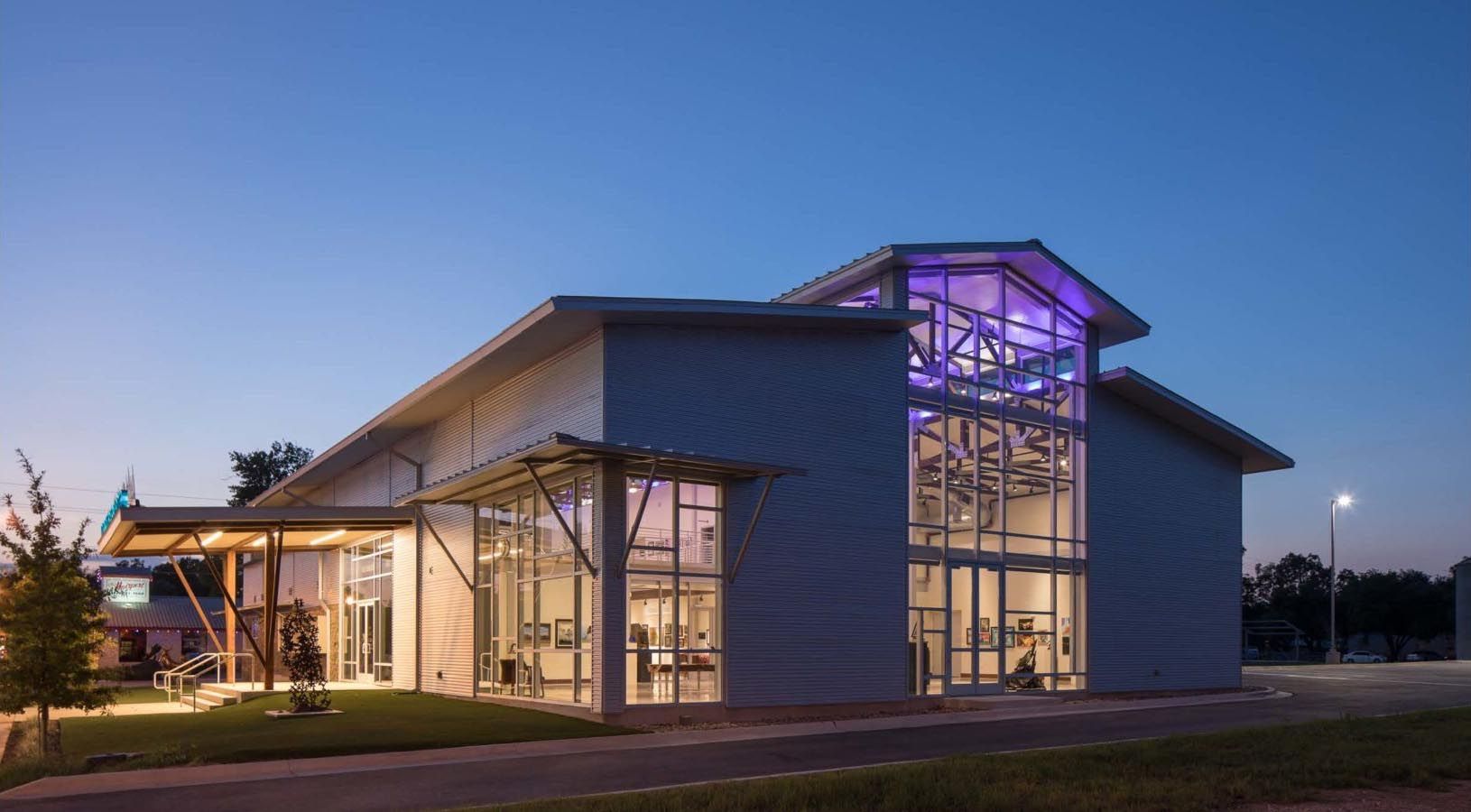 Modern building with glass walls illuminated at dusk. Purple light glows through the upper structure.