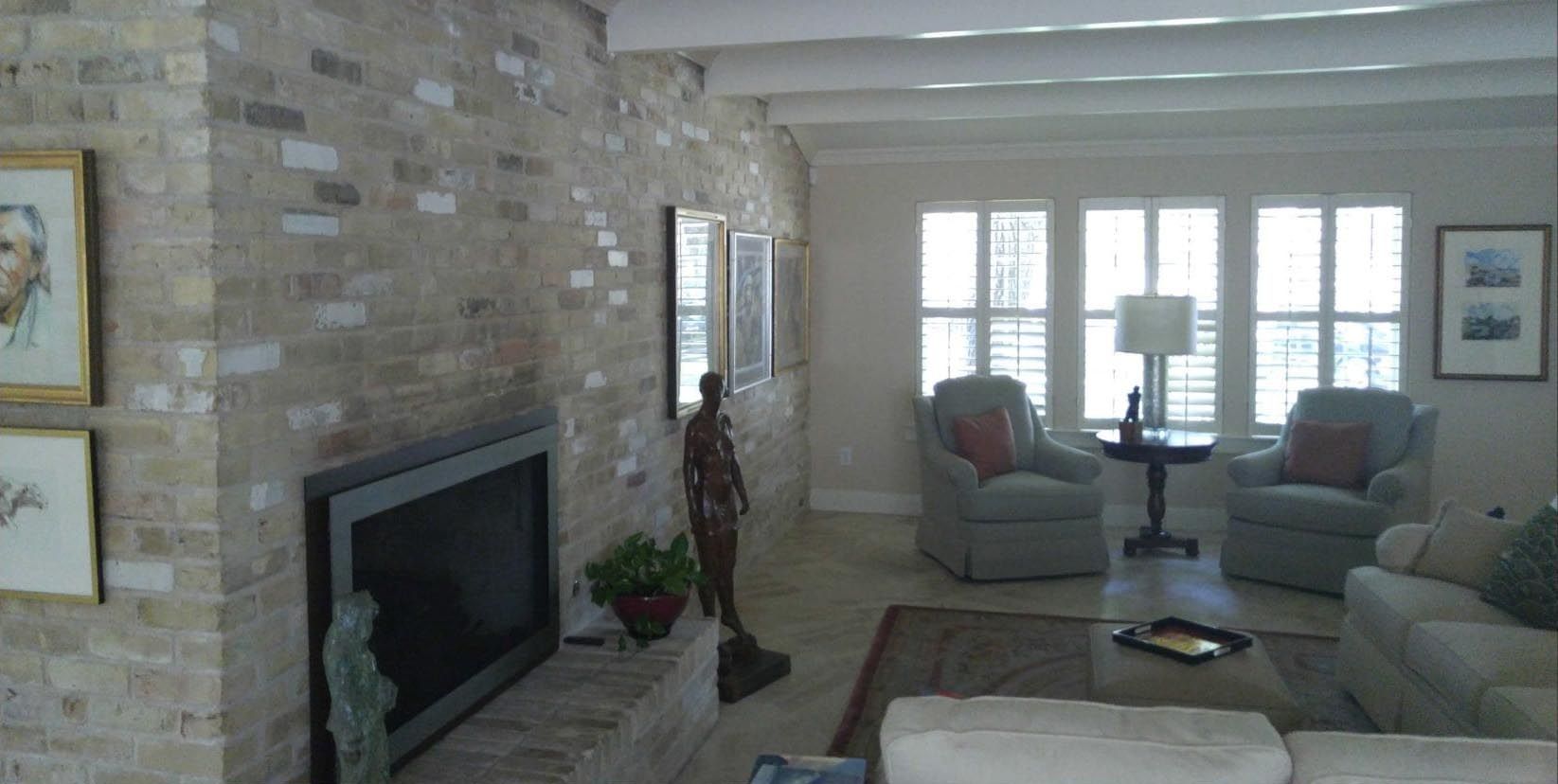 Living room with brick fireplace, TV, chairs, and large window with shutters.