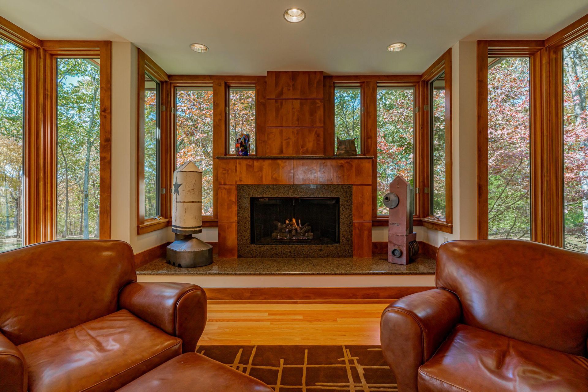 Cozy living room with fireplace, leather armchairs, large windows, and autumn foliage visible outside.