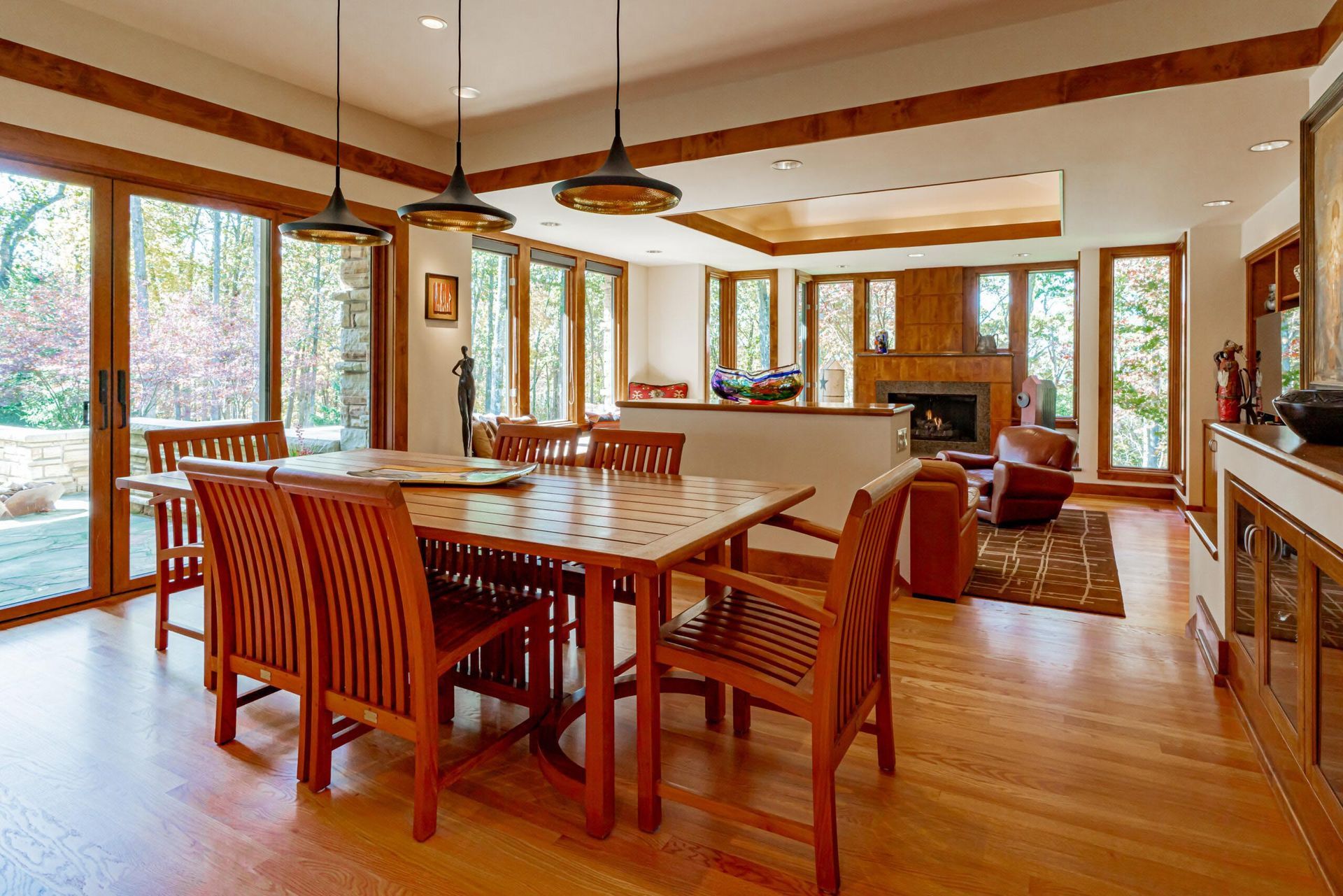 Dining room with wooden table and chairs, large windows overlooking a wooded area.