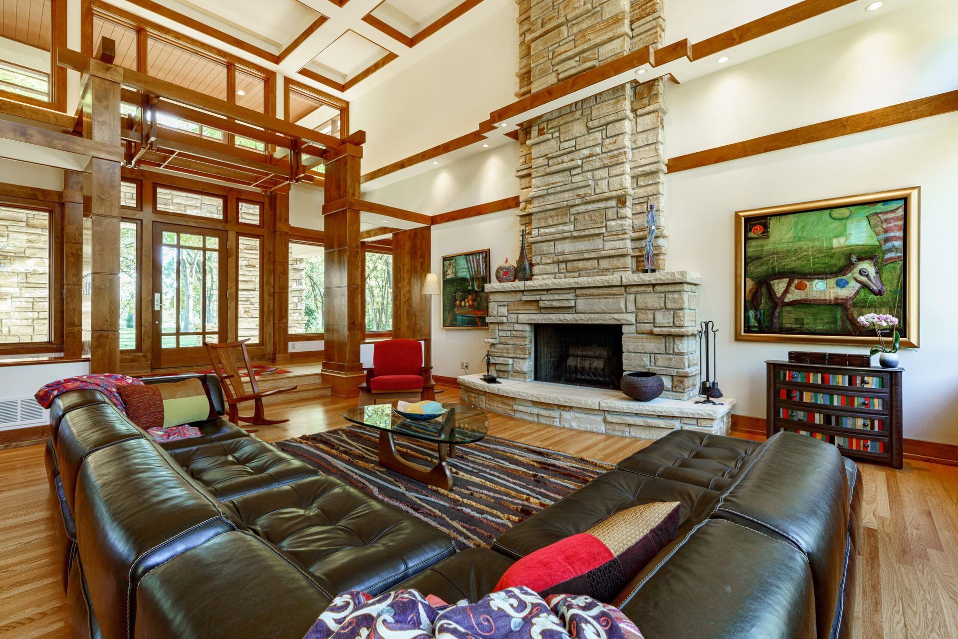 Spacious living room with stone fireplace, leather sectional, artwork, wooden beams, and large windows.