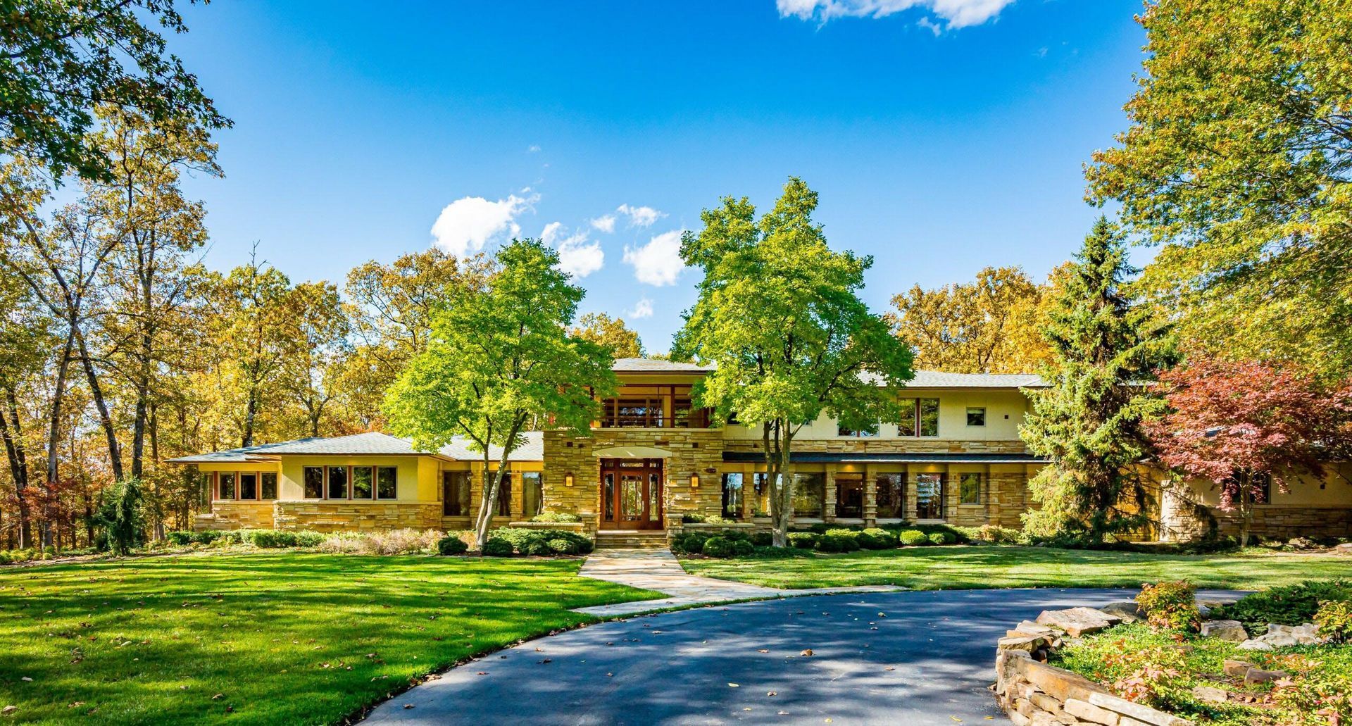 Large, light-colored house with a curved driveway, surrounded by trees with autumn foliage, under a blue sky.