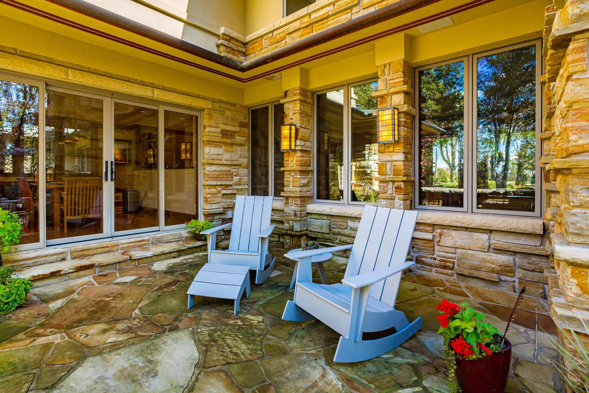 Patio with two blue Adirondack chairs on stone flooring, with views through large windows and a red potted plant.