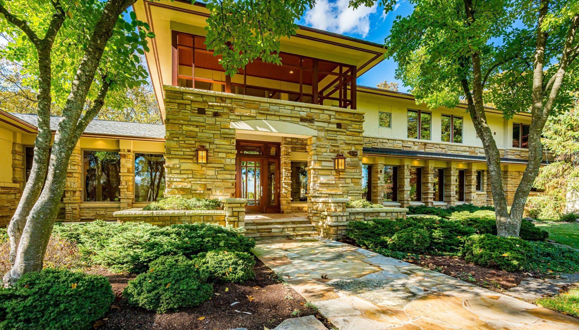 Stone-faced, two-story house with trees and shrubs. Brown door, glass windows, and a covered porch.