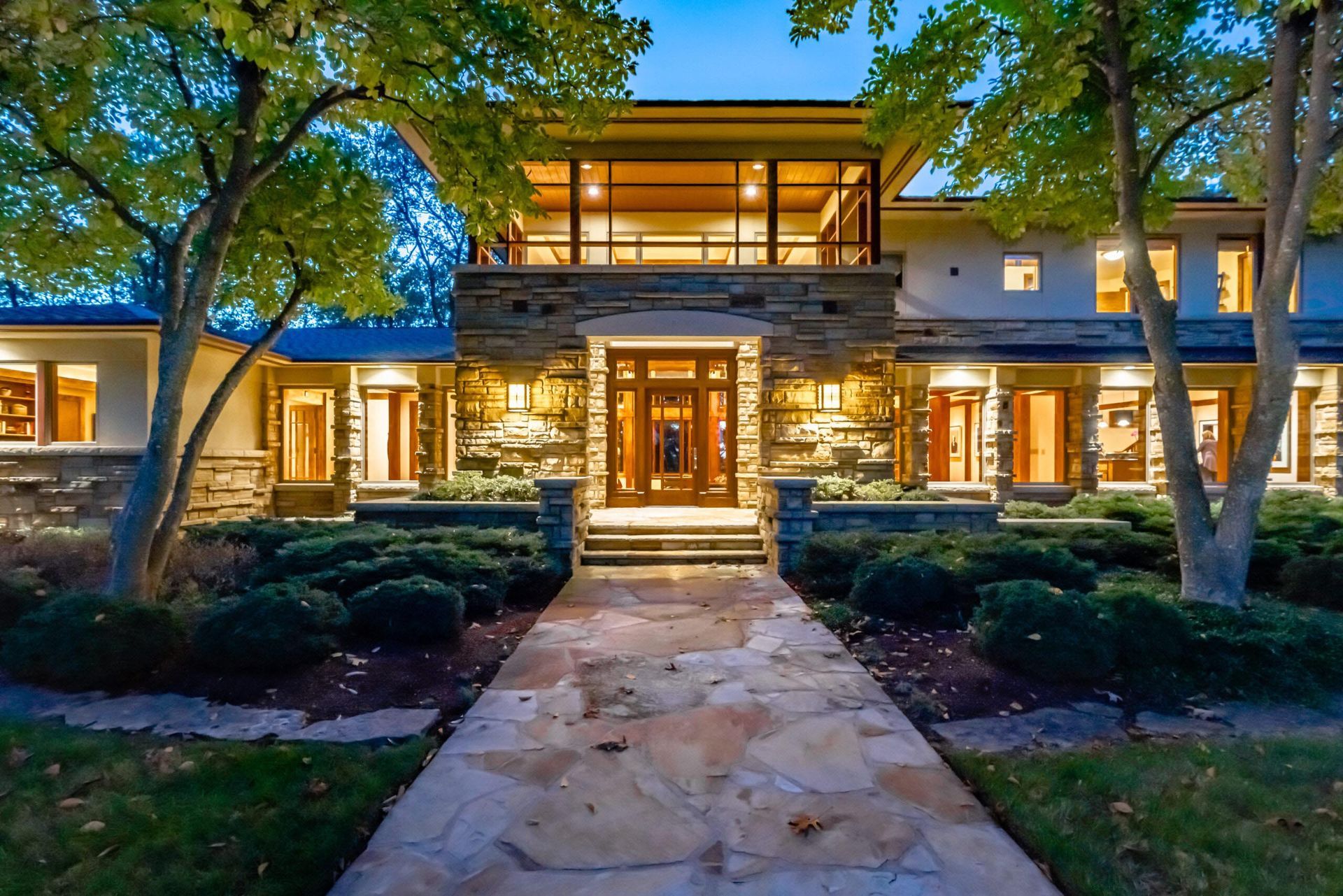 Luxurious house exterior with stone and glass facade, lit windows, stone pathway, and landscaped yard.
