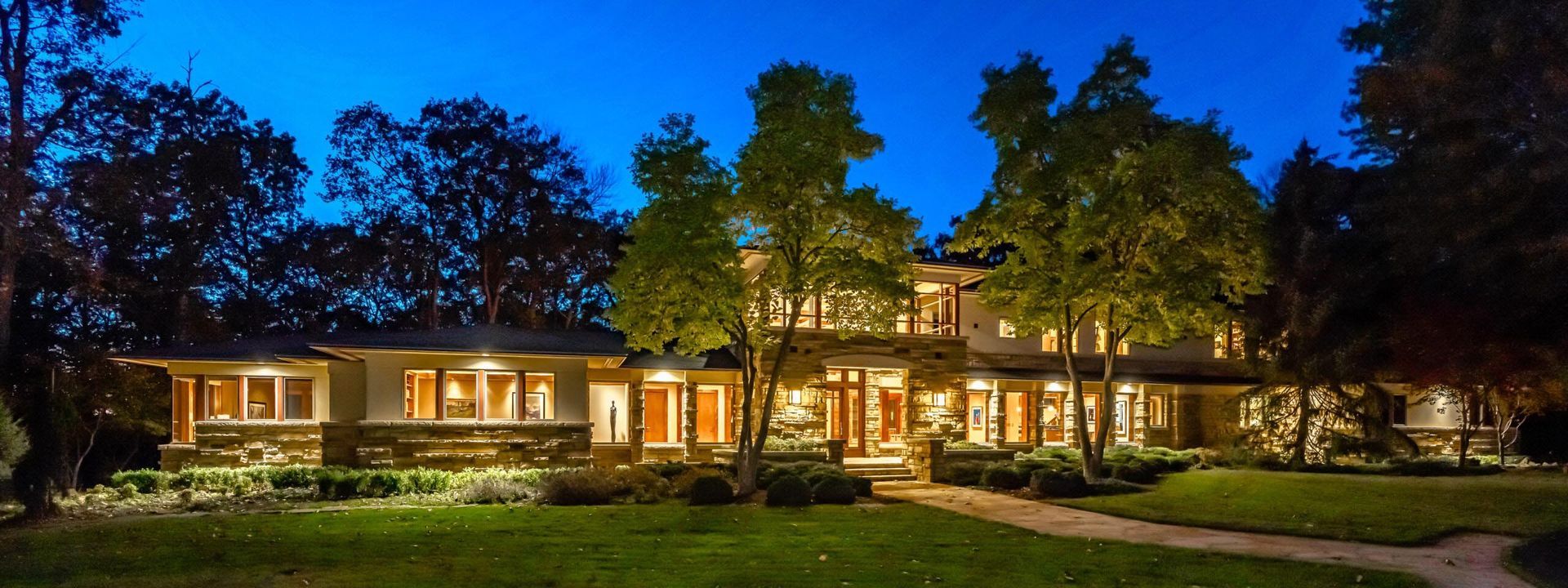 A large, illuminated house at dusk, framed by trees against a blue sky.