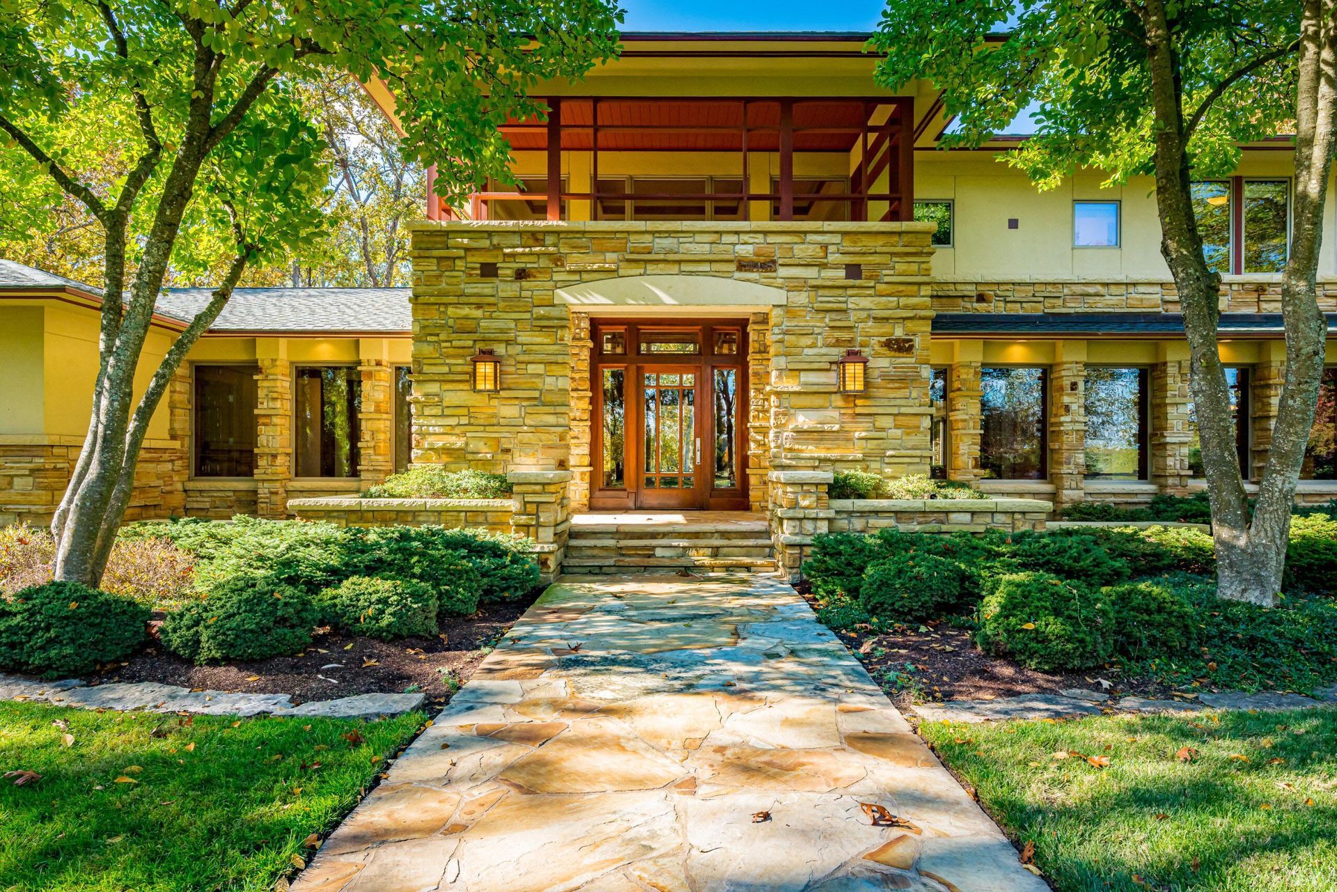 Stone and wood-paneled house with a walkway leading to the front door, flanked by green bushes and trees.