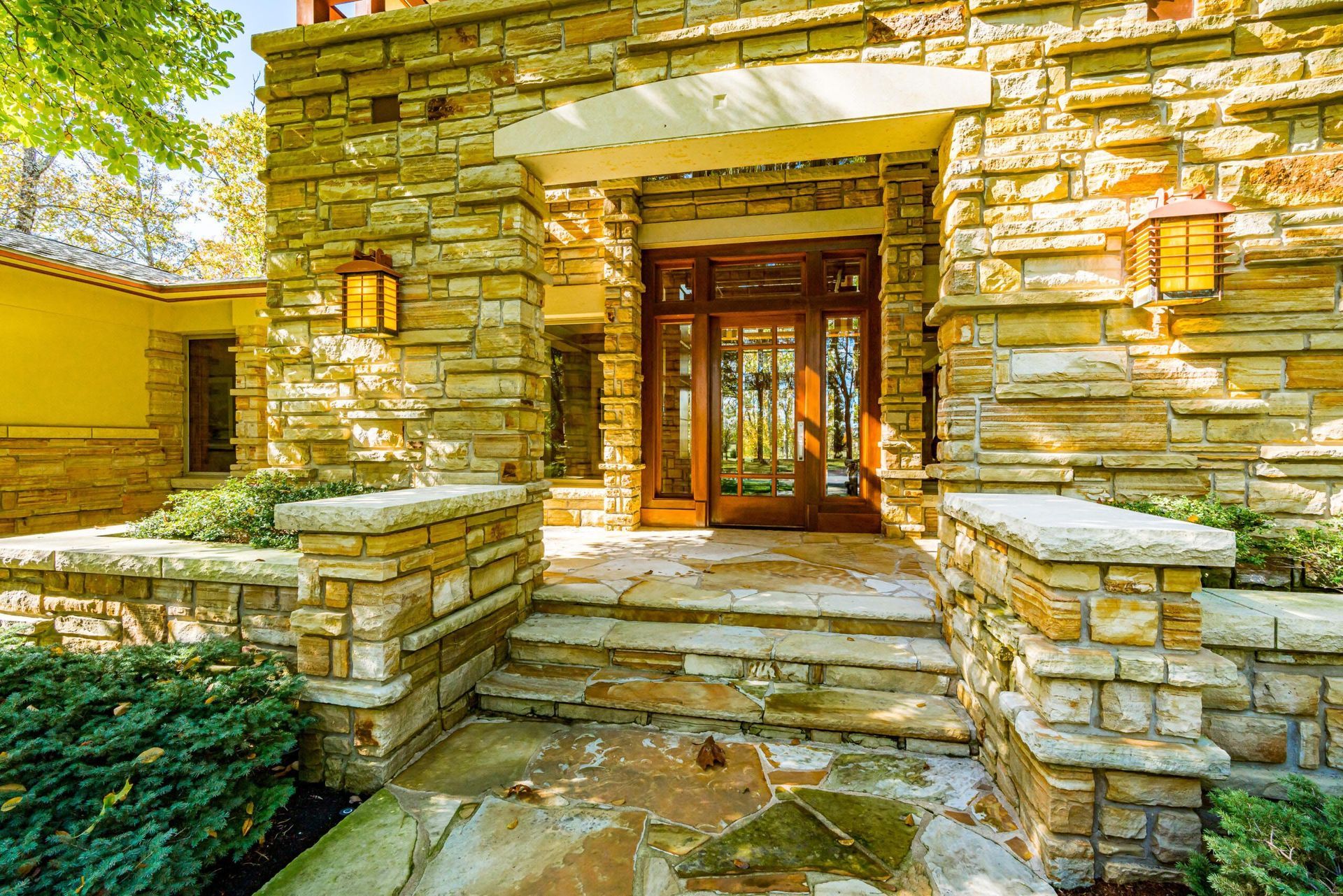 Stone entrance of a house with wooden doors, lit by sconces, stone steps, and surrounding greenery.