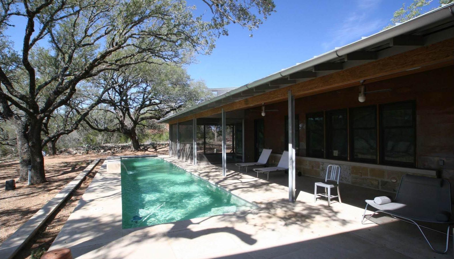 Poolside patio with pool, lounge chairs, and building with windows under a tree.