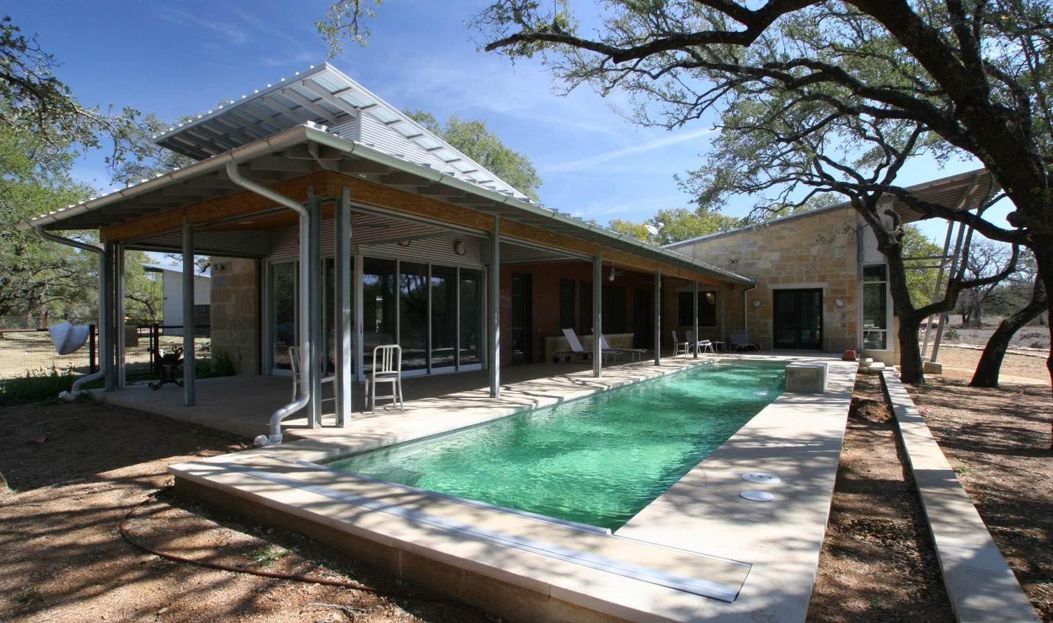 Modern house with pool and covered patio. Stone walls, metal roof, trees in sunny outdoor setting.