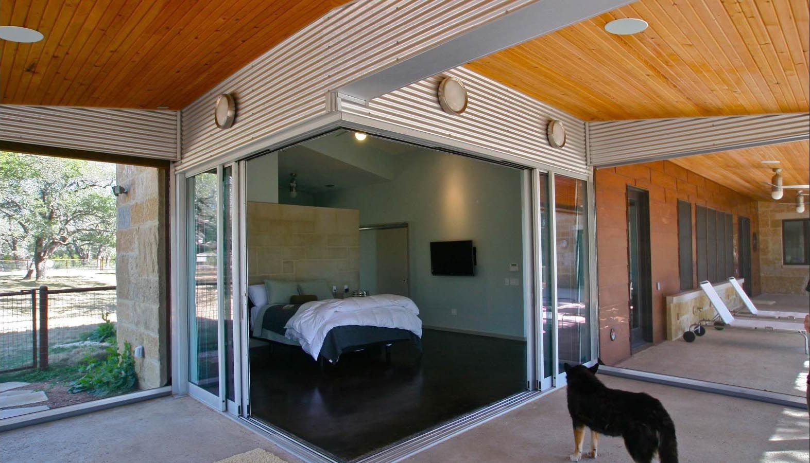Open-air bedroom with glass doors, dog, and patio. Brown ceiling, dark floor, light-colored walls.