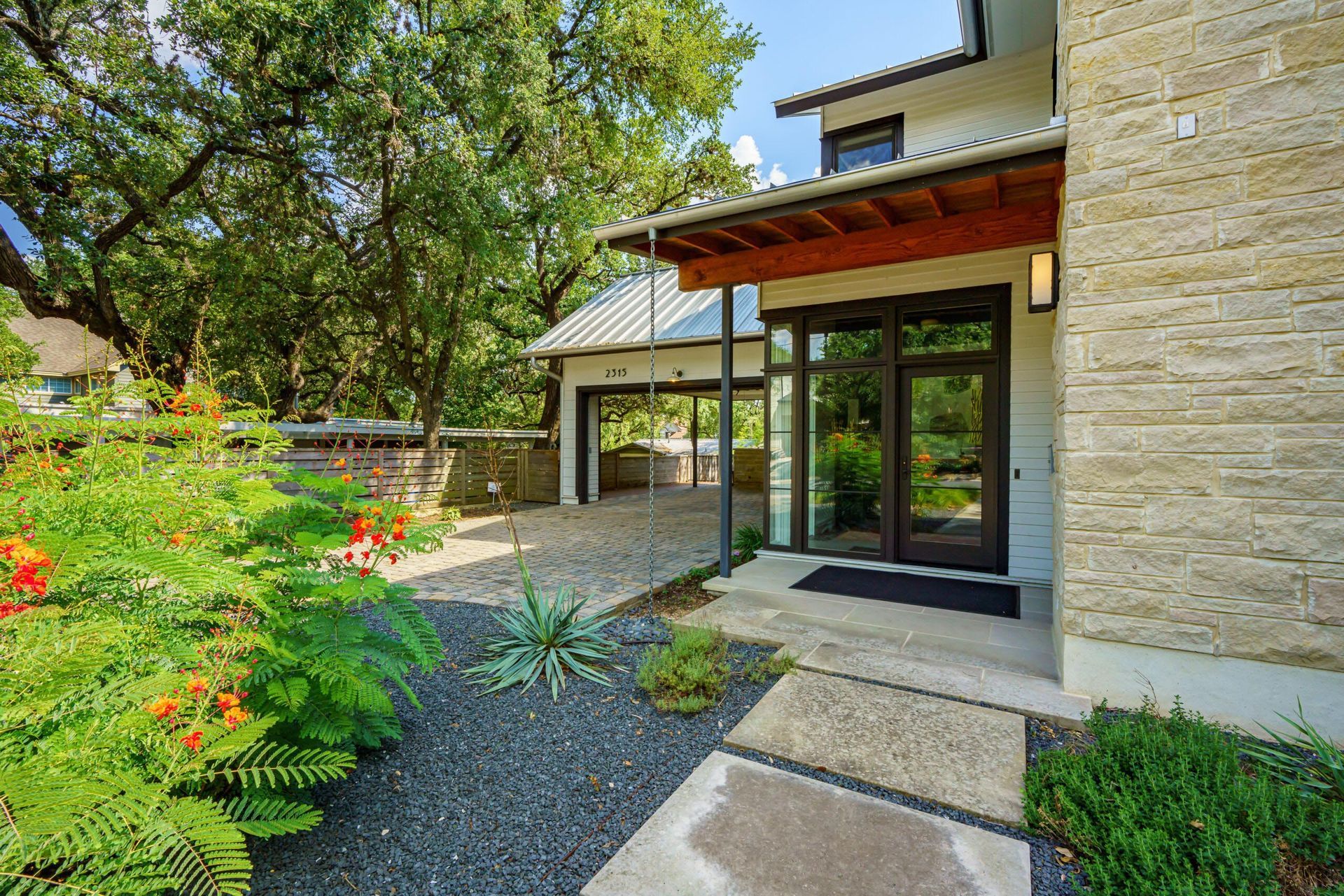 Modern home exterior with stone and glass entry, carport, and lush landscaping.