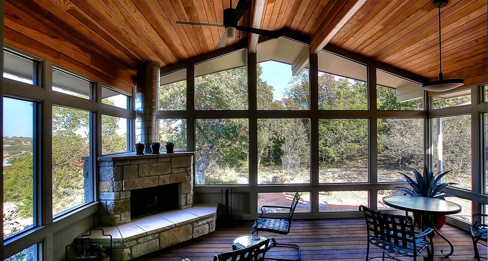 Sunroom with stone fireplace, wood ceiling, large windows overlooking trees.