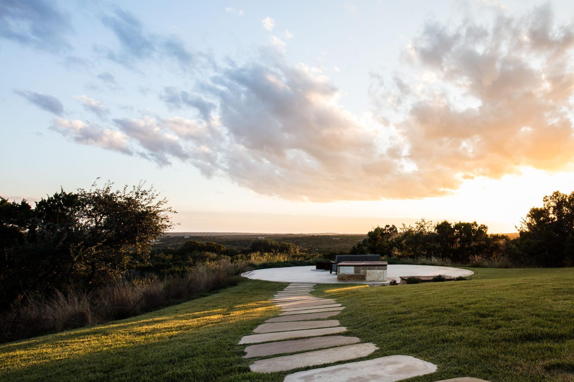 Stone path leads to a circular patio with a small structure, overlooking a landscape at sunset.