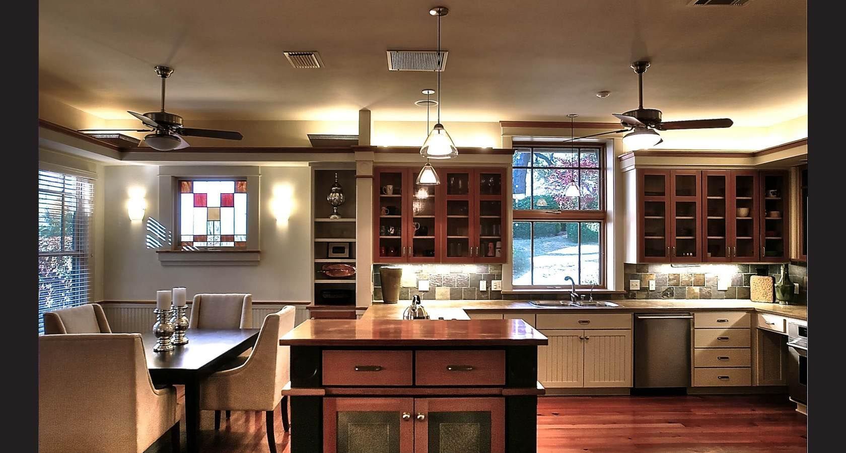 Kitchen interior with wooden cabinets, island, and dining area. Sunlight streams through windows.