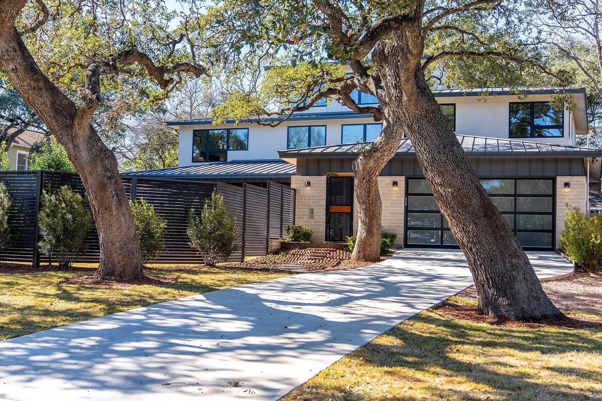 Modern house with a long driveway framed by trees, light-colored facade, and a glass garage door.