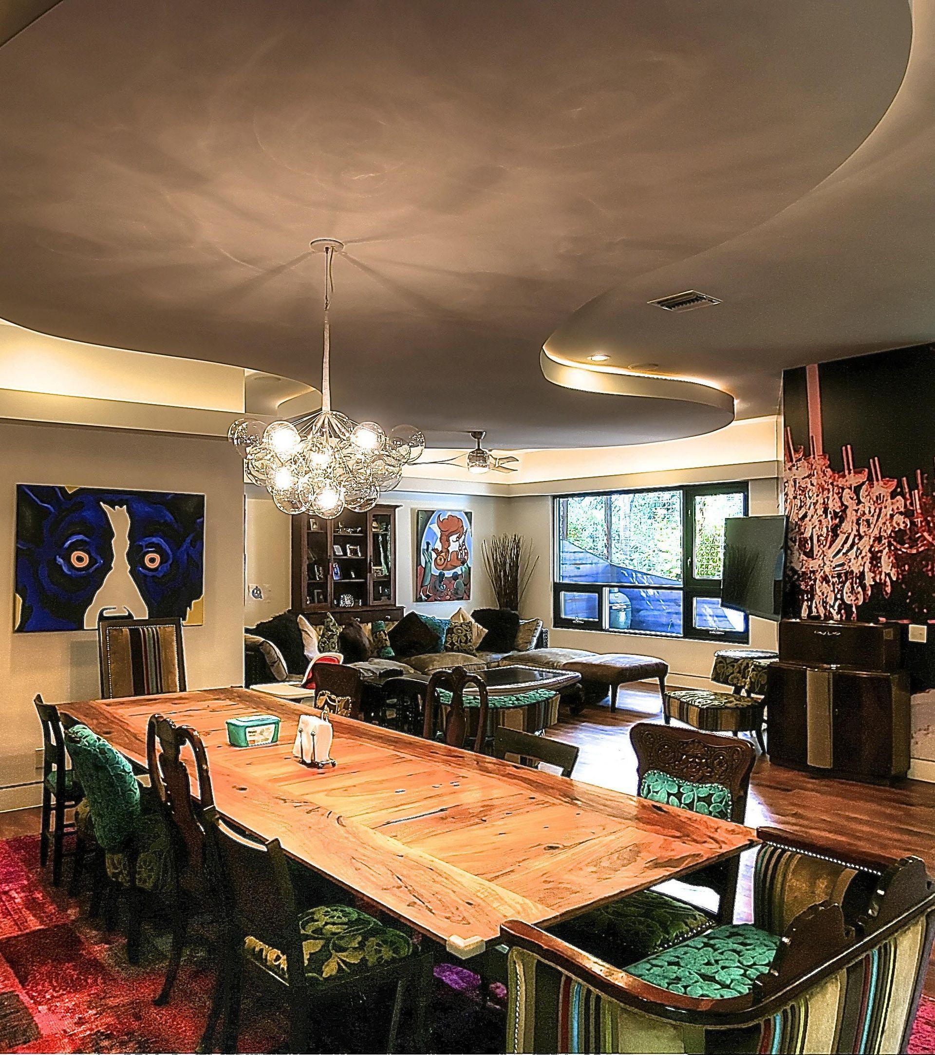 Elegant dining room with large wooden table, chandelier, art, and curved ceiling lighting.