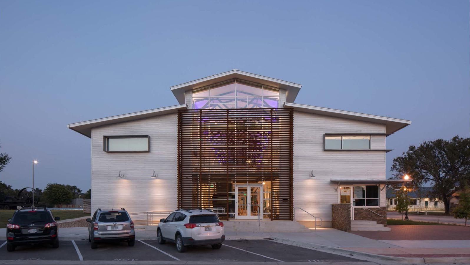 Modern, two-story building with a glass facade, lit up at dusk. Cars parked in front.