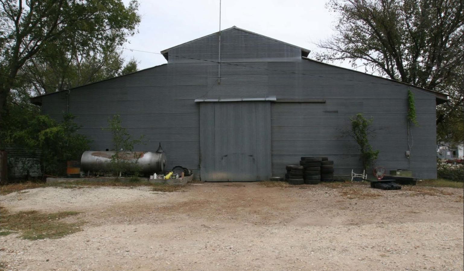 Gray metal barn with a closed sliding door, set on gravel, with trees in the background.