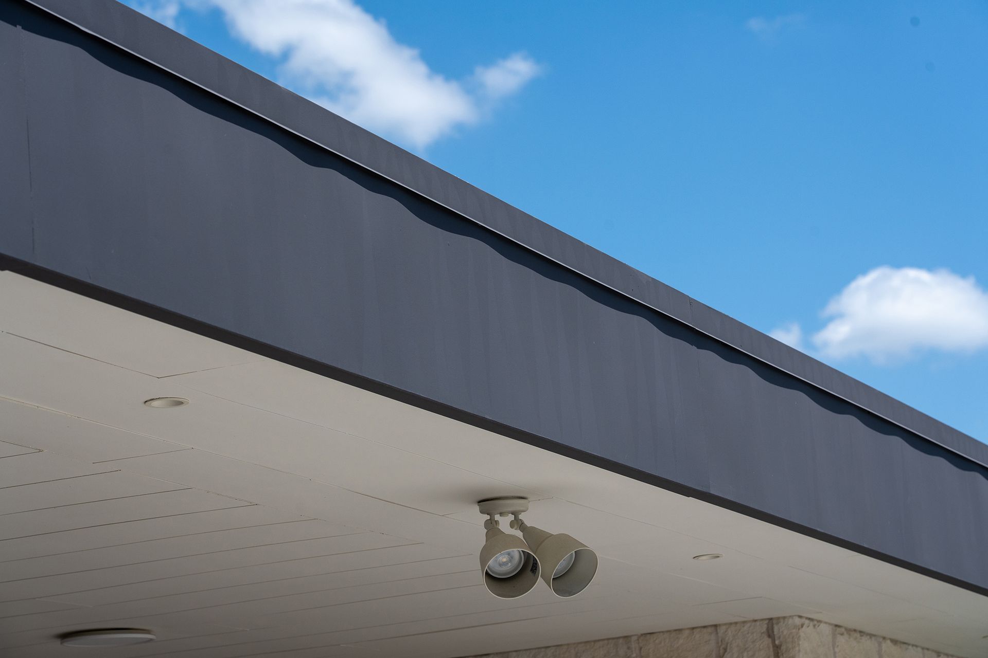 Dark gray building awning with white underside, two light fixtures, blue sky with clouds.