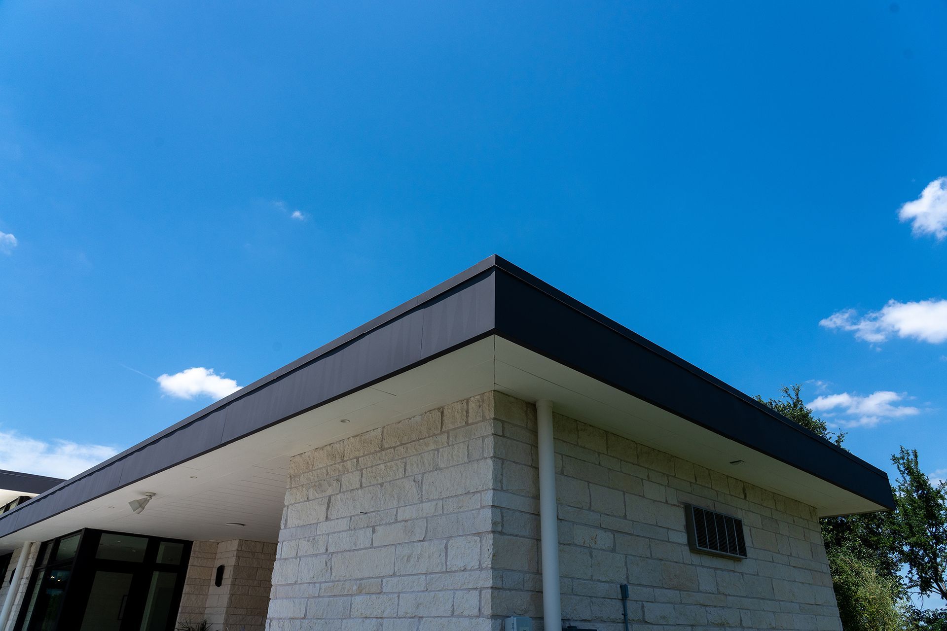 Corner of a modern building with a dark roof against a bright blue sky with clouds.