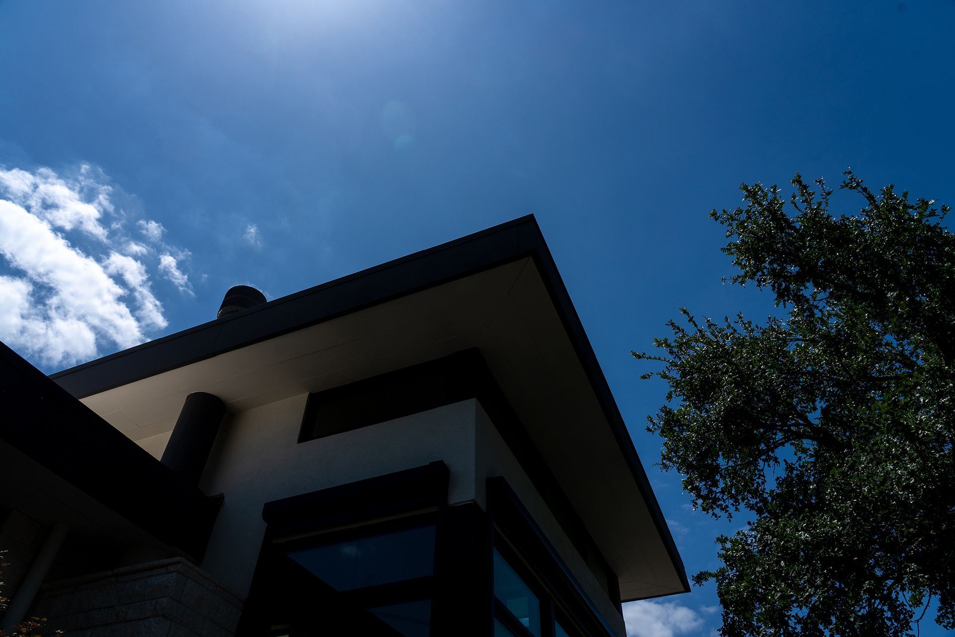 Low-angle view of a modern building against a bright blue sky with white clouds and a tree on the right.