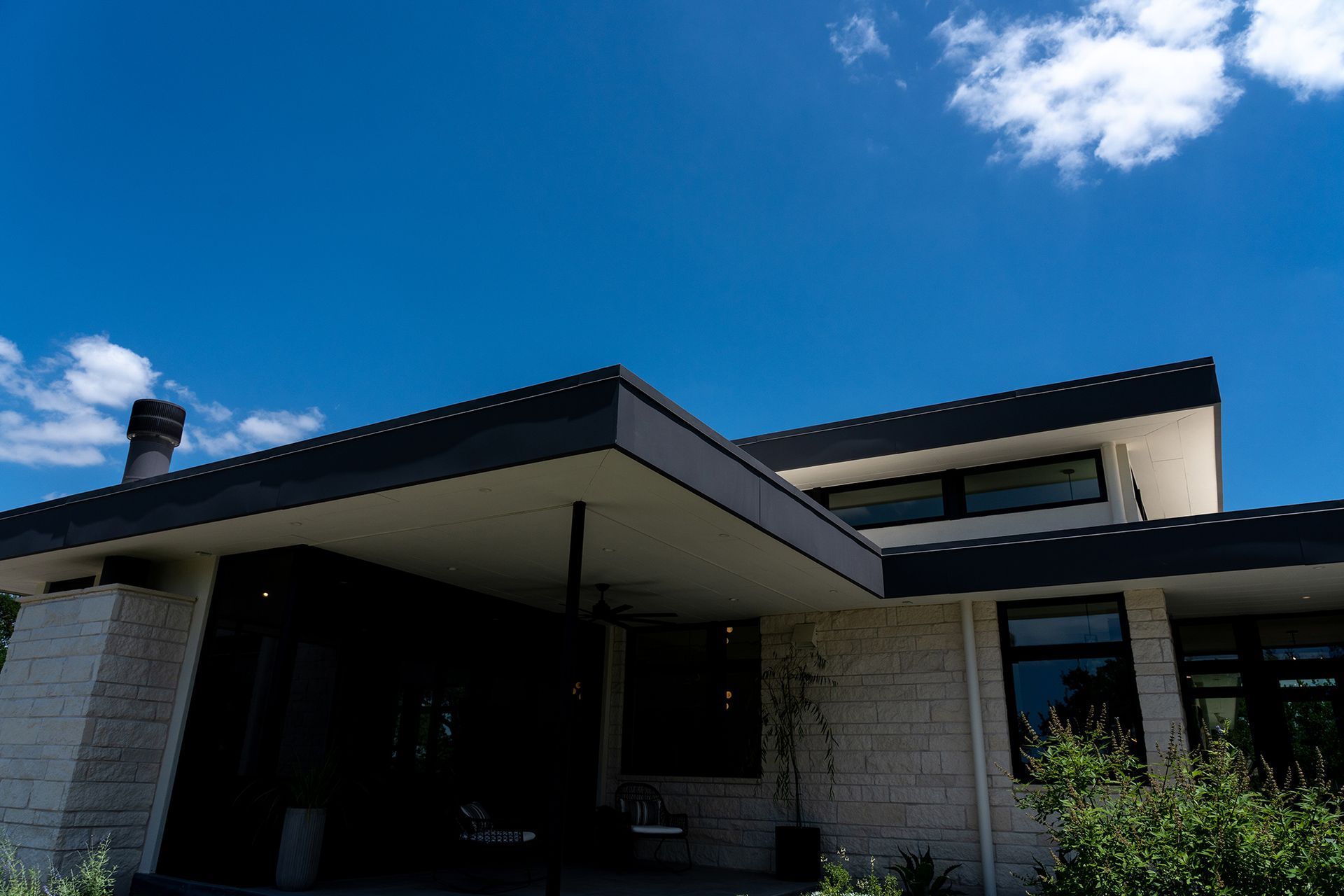 Modern house exterior with flat roof, light stone facade, dark trim, and bright blue sky.