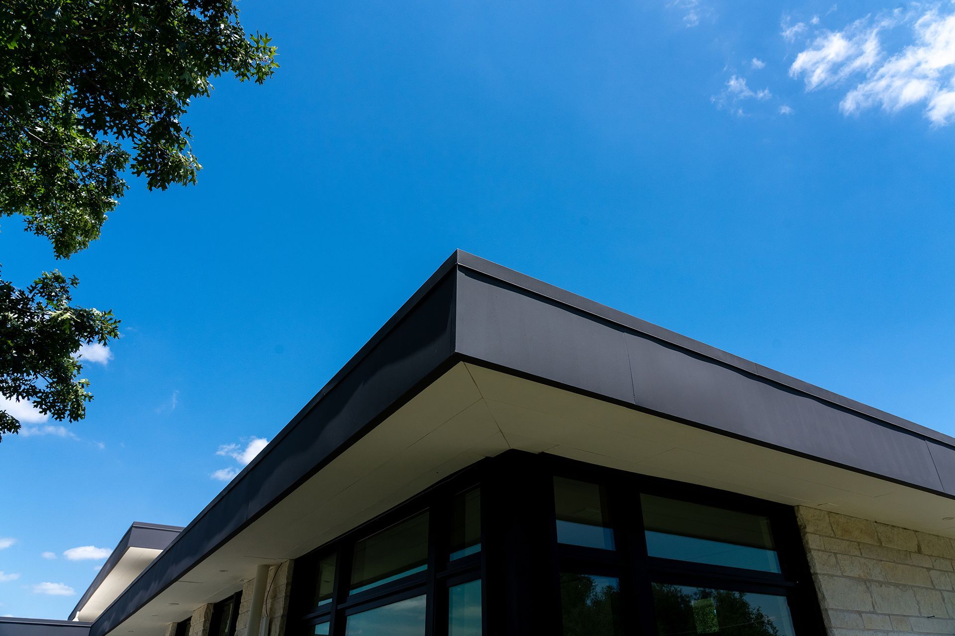 Corner of a modern building with dark trim, below a bright blue sky with a few clouds and tree branches.