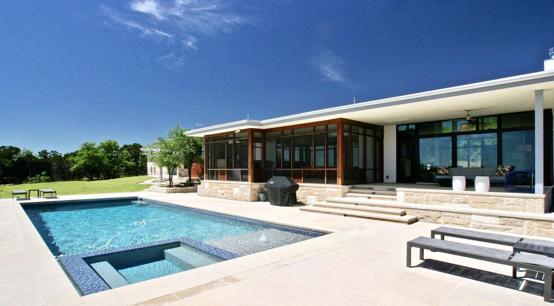 Pool with steps, patio, and modern house under a bright blue sky.