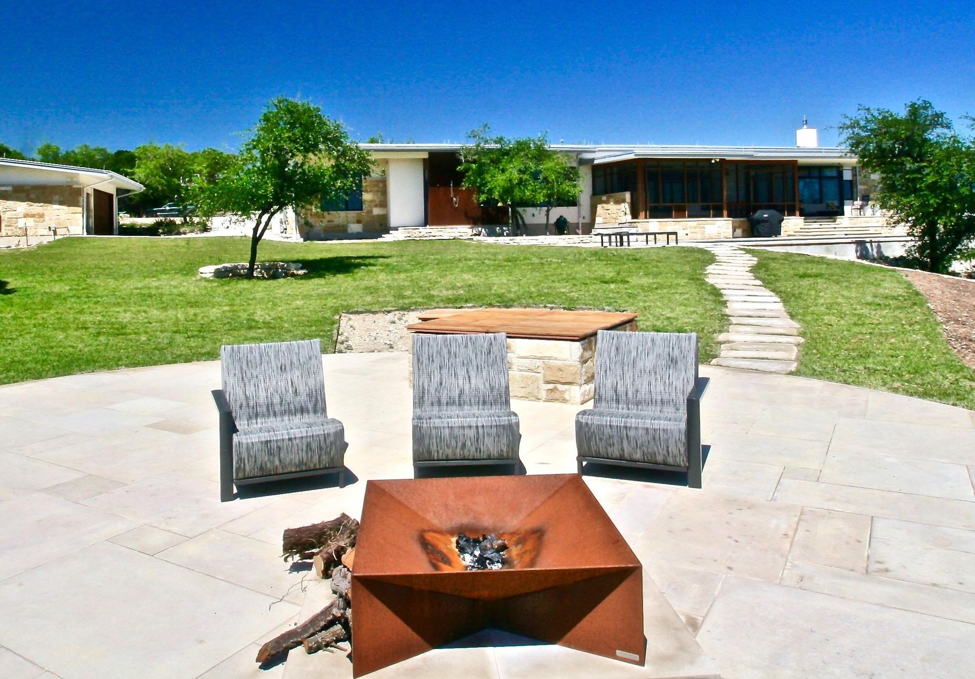 Patio with fire pit, three chairs, and a house in the background on a sunny day.
