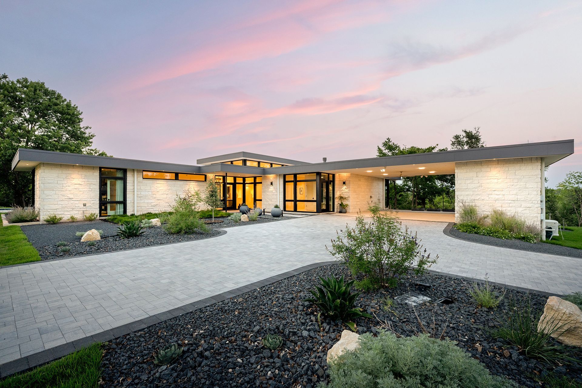 Modern home with a curved roof, gray driveway, and landscaping under a pastel sunset.