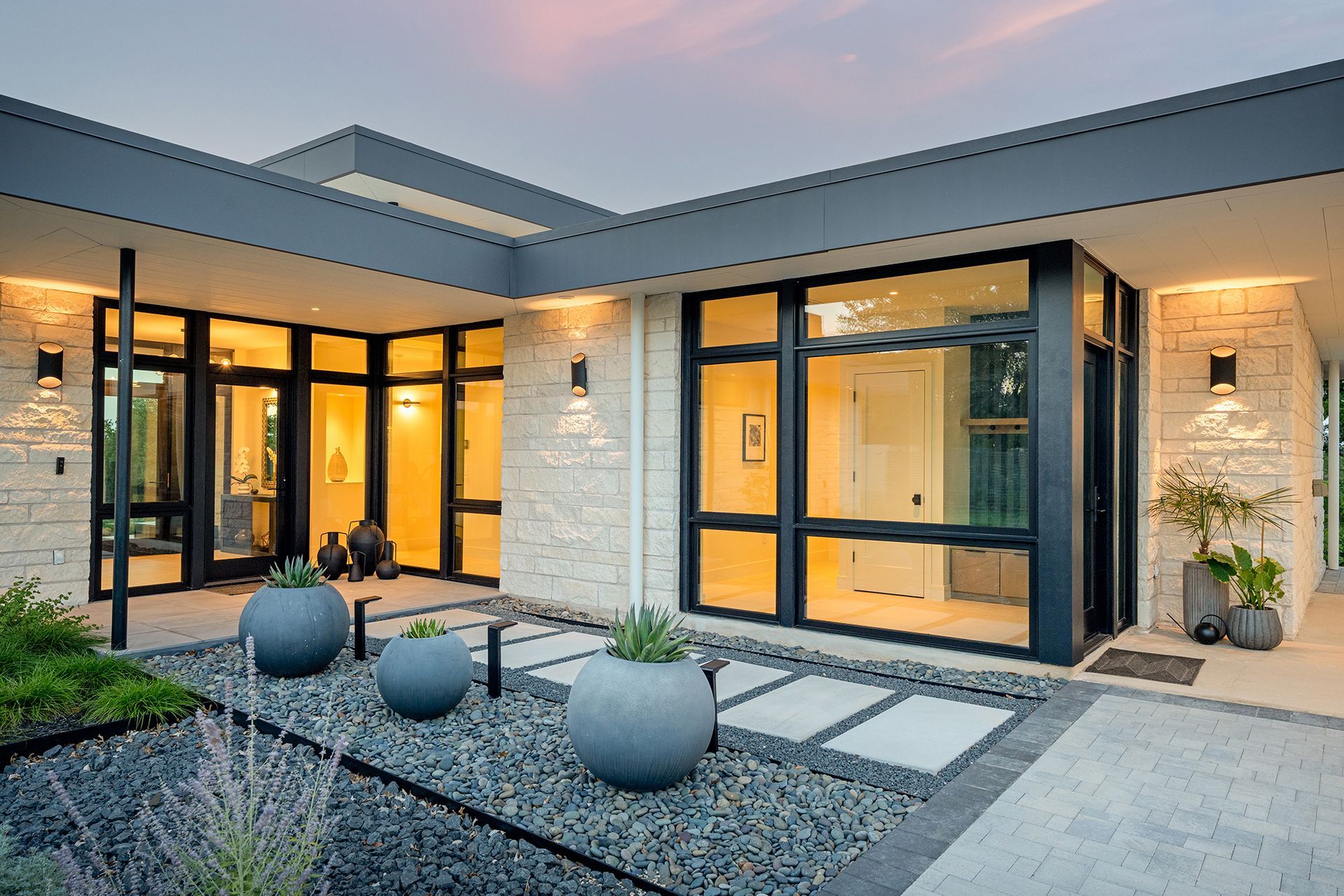 Modern home exterior with large windows, stone facade, and gray trim, at dusk.
