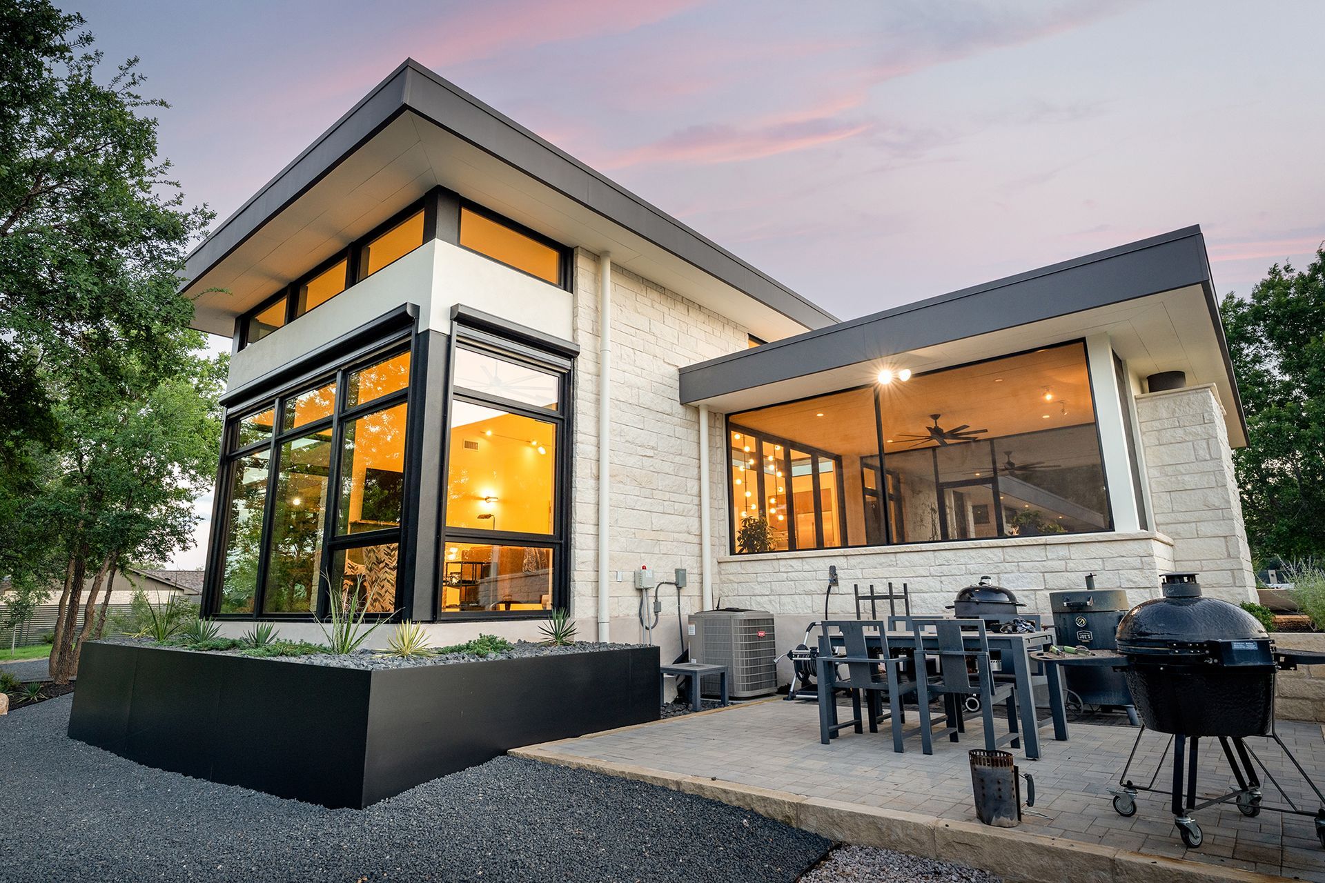 Modern two-story house with large windows, light brick exterior, and outdoor patio with a grill.